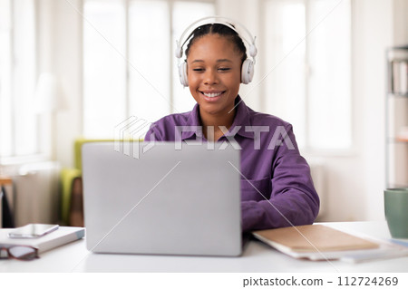 Smiling female black student in headphones using laptop at desk at home 112724269
