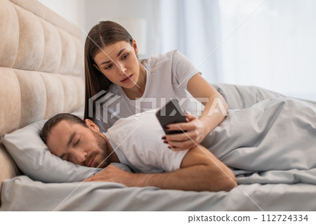 Anxious woman checking phone beside sleeping man Anxious woman checking phone beside sleeping man 112724334