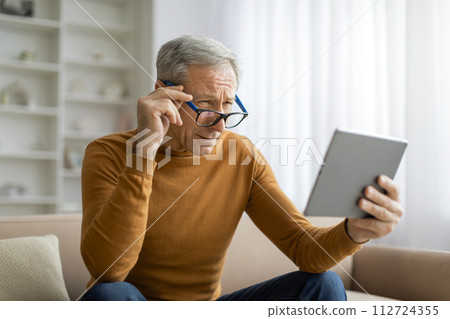 Senior man adjusting his eyeglasses while using tablet at home Senior man adjusting his eyeglasses while using tablet at home 112724355