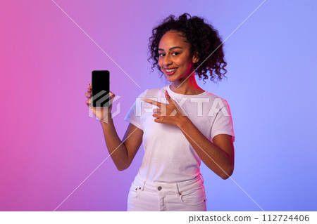 Cheerful black woman pointing at smartphone with blank screen on neon background 112724406