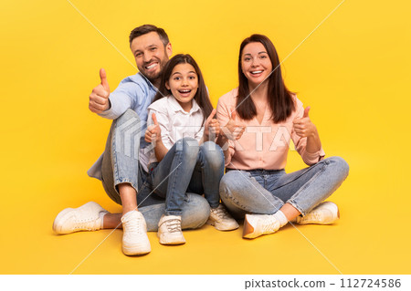 Happy family of three giving thumbs up sitting, yellow backdrop Happy family of three giving thumbs up sitting, yellow backdrop 112724586