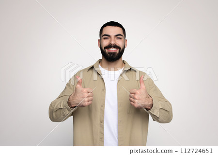 Optimistic bearded man in a beige shirt and white t-shirt giving two thumbs up with a broad smile 112724651