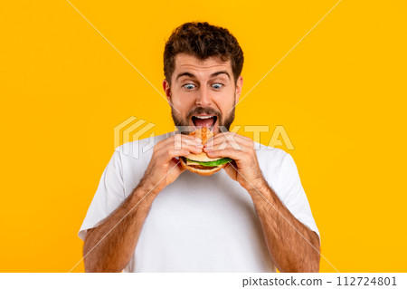 Excited hungry Caucasian man biting delicious cheeseburger against yellow backdrop Excited hungry Caucasian man biting delicious cheeseburger against yellow backdrop 112724801