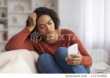 Distressed black woman sitting on couch and looking at smartphone with anxiety 112724802