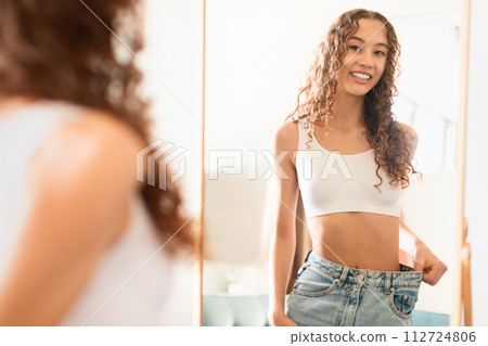 Caucasian teenager girl at mirror showcasing weight loss success indoors Caucasian teenager girl at mirror showcasing weight loss success indoors 112724806