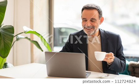 Joyful mature man with a beard working on a laptop and enjoying a cup of coffee in a bright office Joyful mature man with a beard working on a laptop and enjoying a cup of coffee in a bright office 112724824