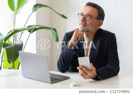 Pensive senior man with glasses holding a smartphone and looking thoughtfully into the distance 112724902