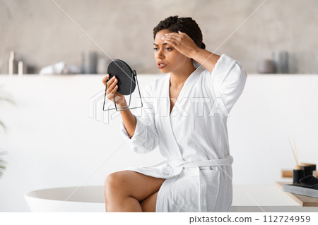 Young black woman in white bathrobe holding mirror, critically examining her skin Young black woman in white bathrobe holding mirror, critically examining her skin 112724959