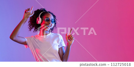 Joyful black woman with sunglasses and headphones dancing on neon background, free space 112725018