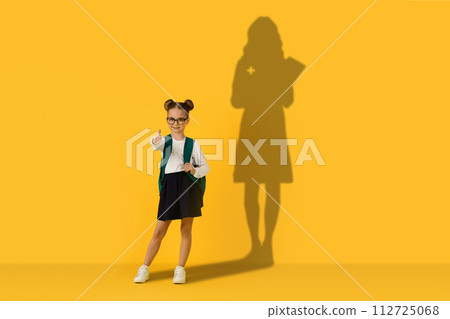 A confident schoolgirl with glasses and hair buns gives a thumbs up while holding a green backpack 112725068