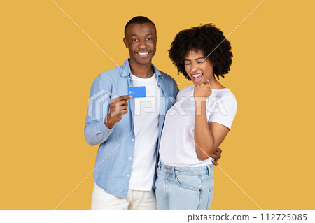 Confident young man showcasing a credit card with a proud smile while the delighted woman cheers 112725085