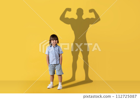 A cheerful young child stands smiling with a confident, muscular superhero shadow cast 112725087