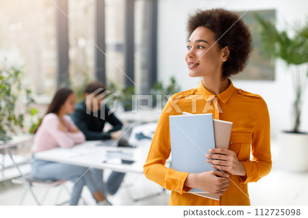 Confident black lady student holding books with classmates in background 112725098