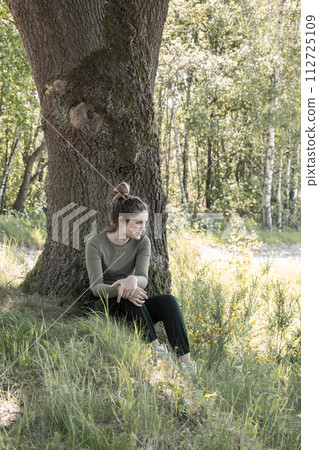 Relaxed Young Woman Sitting Under Tree in Sun-Dappled Woods Relaxed Young Woman Sitting Under Tree in Sun-Dappled Woods 112725109