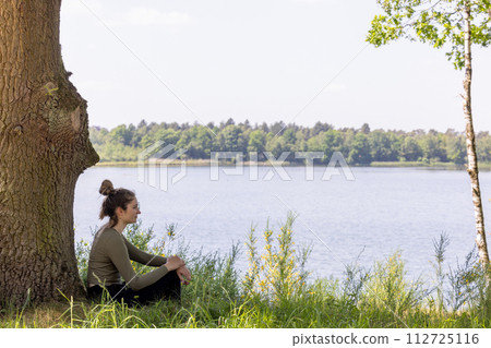 Tranquil Respite: Young Woman Resting Against Tree by the Lake 112725116