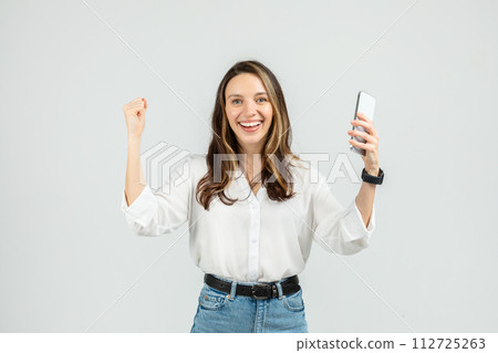 Exuberant young woman with a smartwatch, triumphantly raising one fist while holding a smartphone 112725263