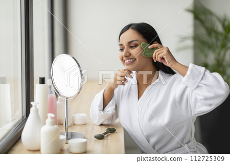Smiling indian woman using gua sha, looking at mirror 112725309