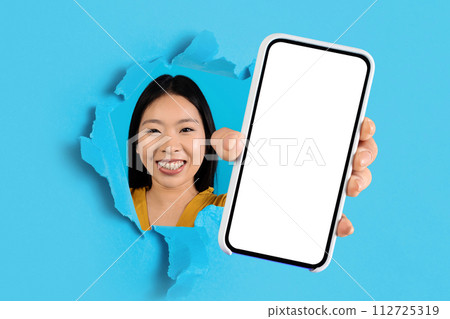 Cheerful young Asian woman smiling through a torn blue paper, presenting a smartphone with a blank screen Cheerful young Asian woman smiling through a torn blue paper, presenting a smartphone with a blank screen 112725319