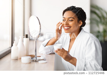 Smiling black woman in bathrobe applying cream to her cheek 112725446
