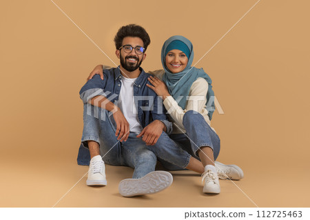 Stylish young muslim couple in casual denim outfits sitting on beige background Stylish young muslim couple in casual denim outfits sitting on beige background 112725463