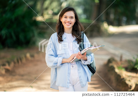 A cheerful young female student with a bright smile walks on a forested campus path 112725464