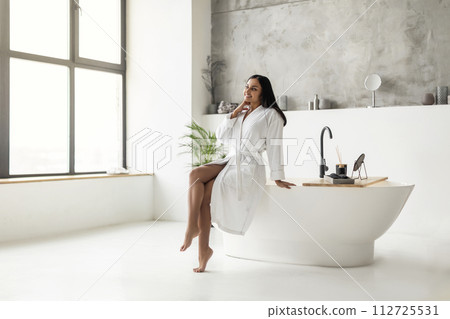 Cheerful young eastern woman in bathrobe sitting on bathtub 112725531