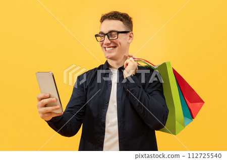 Cheerful young man with purchases and smartphone, yellow background Cheerful young man with purchases and smartphone, yellow background 112725540