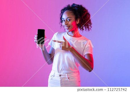 Happy black lady pointing to smartphone on vibrant pink and purple backdrop 112725742
