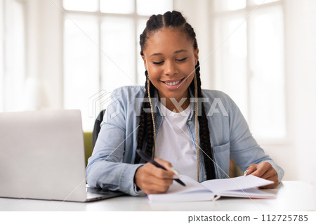 Happy african american female teen studying with laptop and notebook at home 112725785