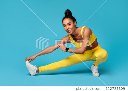 Black sportswoman in yellow fitwear stretching legs against blue background 112725809