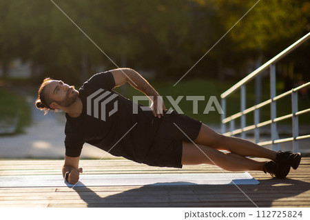 Muscular guy in sportswear doing side elbow plank exercise outside 112725824