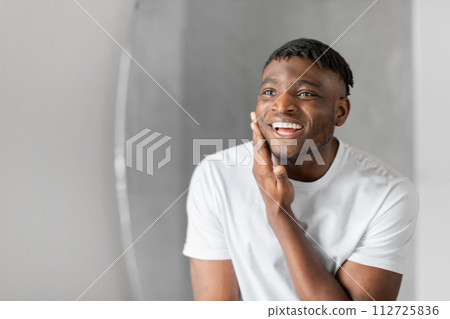black guy stands in bathroom looking at mirror touching face 112725836