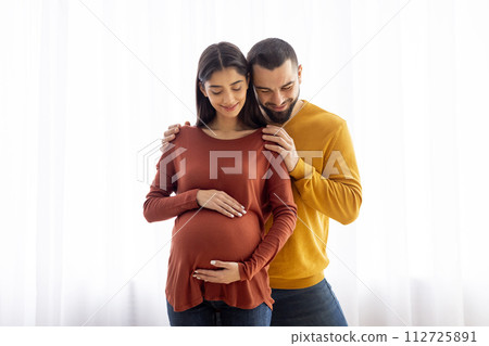 Pregnant woman and her husband sharing tender embrace near window at home Pregnant woman and her husband sharing tender embrace near window at home 112725891