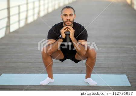Young sporty man in black fitwear squatting holding dumbbell outdoor Young sporty man in black fitwear squatting holding dumbbell outdoor 112725937