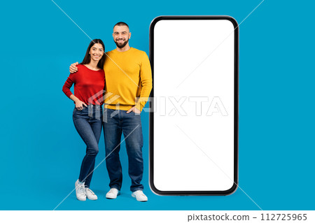 Happy young couple standing next to giant smartphone with blank white screen Happy young couple standing next to giant smartphone with blank white screen 112725965