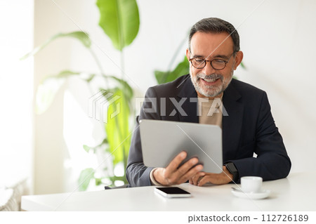 Cheerful mature man in a smart suit and turtleneck, engaging with a digital tablet 112726189