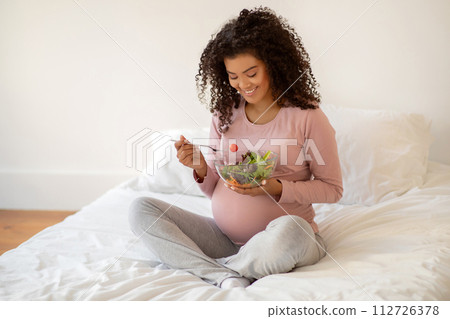 Beautiful black pregnant woman sitting on bed and holding bowl of salad Beautiful black pregnant woman sitting on bed and holding bowl of salad 112726378