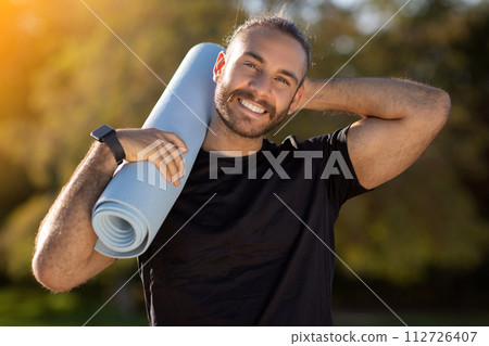 Cheerful and fit European man holding fitness mat stands outdoors 112726407