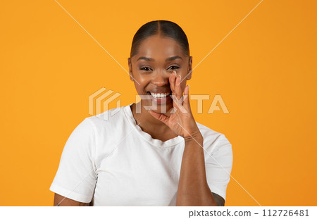 Cheerful African American woman stands against orange backdrop sharing secret 112726481