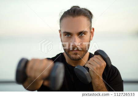 Determined man in fitwear exercises with dumbbells flexing biceps outside Determined man in fitwear exercises with dumbbells flexing biceps outside 112726533