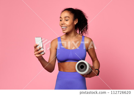 Athletic African woman holds smartphone and foam roller, pink background 112726620