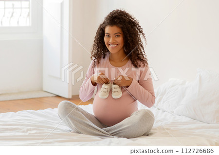 Beautiful black pregnant woman sitting on bed and holding tiny baby shoes 112726686