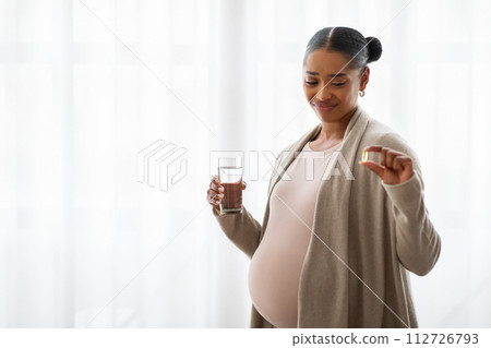 Unhappy pregnant black woman showing capsule, holding glass of water 112726793