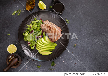 grilled cooked piece of tuna fillet with sliced avocado and salad on black ceramic plate 112726794