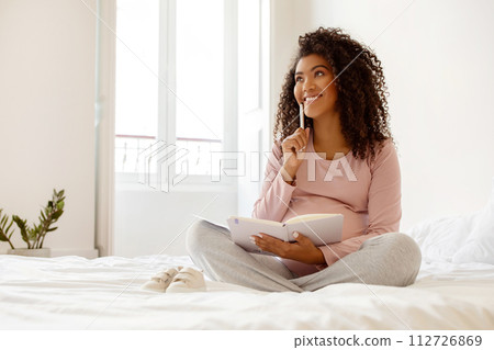 Happy Black Pregnant Woman Writing Checklist In Notepad While Sitting On Bed 112726869