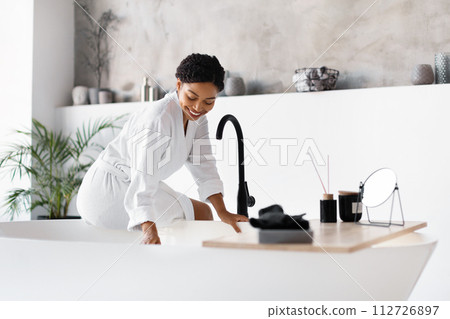 Smiling black woman in bathrobe leaning over modern freestanding bathtub at home Smiling black woman in bathrobe leaning over modern freestanding bathtub at home 112726897