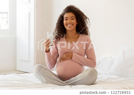 Smiling black pregnant woman sitting on bed and holding glass of milk 112726931