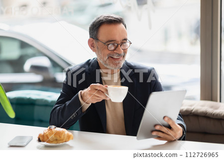 Senior man in a smart casual outfit enjoying a cup of coffee while reading on a tablet in a cafe 112726965