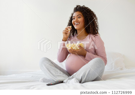 Joyful pregnant black woman eating popcorn while sitting on bed at home Joyful pregnant black woman eating popcorn while sitting on bed at home 112726999
