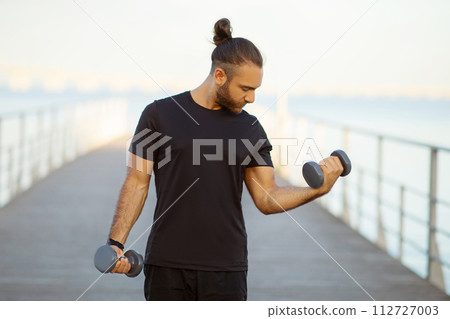 Strong bodybuilder guy flexing his biceps exercising with dumbbells outdoors Strong bodybuilder guy flexing his biceps exercising with dumbbells outdoors 112727003